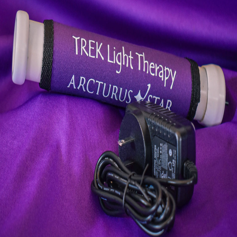A purple cylindrical light therapy device labeled 'TREK Light Therapy ARCTURUS STAR' with white caps on both ends, shown with a black power adapter and cord on a purple fabric background.