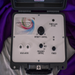 Lymphstar Essence Lymphatic Therapy device in a black carrying case, showing the front control panel with knobs, jacks, and a printed logo on a white background with purple fabric draped behind it.