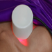 A white circular handheld device emitting red light onto a person's skin, with purple fabric draped in the background.